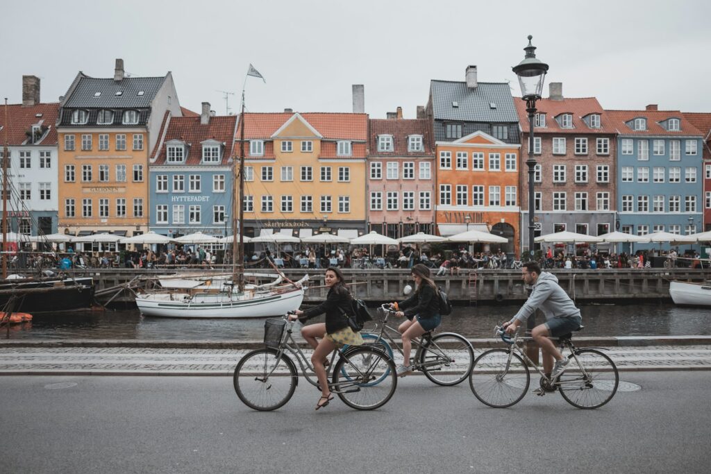 Des jeunes à vélo dans le centre de Copenhague, avec ses bateaux et ses maisons colorés en arrière plan. 